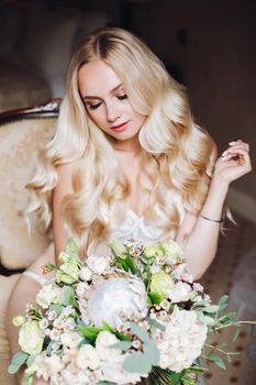 Portrait of beautiful blonde with attractive body, wearing in elegant white lace lingerie looking downn on flowers. Pretty bride holding wedding bouquet in one hand and posing with closed eyes.