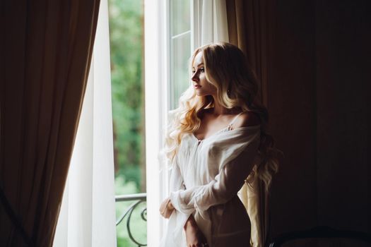 View of gorgeous woman standing near balcony and posing in white lace lingerie. Girl in vintage interior room, looking in big window. Bride at morning.