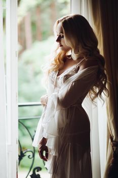 View of gorgeous woman standing near balcony and posing in white lace lingerie. Girl in vintage interior room, looking in big window. Bride at morning.