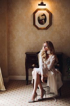 Gorgeous and beautiful blondie bride with long wavy hair, sitting on chair in luxury interior, looking at camera. Seductive woman wearing in white lace lingerie in morning. Wedding concept.