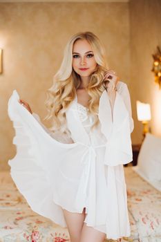 Sexy blondie bride in white lace lingerie and dress posing at luxury bedroom interior and undressing. Hot blonde haired woman holding dress by hand, looking away. Morrning of bride before wedding.