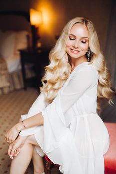 Beautiful and sensuality blondie bride sitting in bedroom, dreaming with closed eyes. Young and sexy woman with long curly hair and perfect make up, wearing in lace lingerie. Morning before wedding.