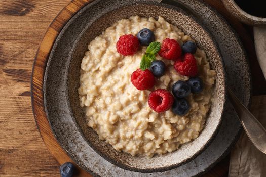 Oatmeal rustic porridge with blueberry, raspberries in ceramic vintage bowl, dash diet with berries, wooden dark brown background, top view close up. Healthy diet breakfast