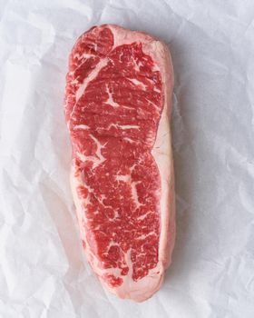 Big whole piece of raw beef meat, striploin on white parchment paper on craft background, zero waste packing. Steak without seasoning and salt. Top view, vertical