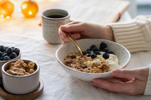 Reading book and eating healthy Christmas holiday winter breakfast with granola muesli and yogurt in bowl on white table background, lights garlands in bokeh. Organic morning diet meal with oat