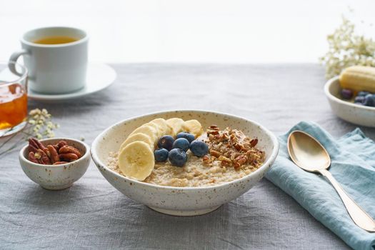 Whole oatmeal, large bowl of porridge with banana, blueberries, nuts for breakfast, morning meal. Side view, close up, gray table. Vegan tasty healthy diet.