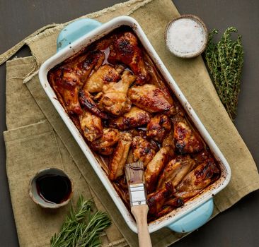 Barbecue chicken wings. Slow cooker sweet and spicy. Oven baked marinated chiken in blue ceramic tray with brush in bbq red sauce. Dark rustic table. Top view, close up