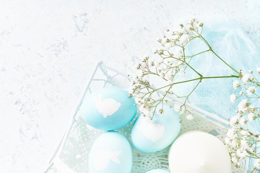 Easter. Holiday. Light white background, gentle pastel colors. Blue eggs with image of rabbit in basket. Flowers in background, top view, copy space,