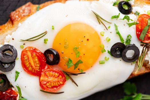Toasted bread toast with fried eggs with yellow yolk and tomatoes, olives, sprinkled with herbs on dark wooden serving board on blue denim napkin, top view, macro, close up