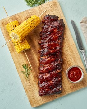 Barbecue pork ribs. Slow cooking recipe. Whole pickled roasted pork meat with red sauce and corn on cutting board, vertical, top view. Turquoise background
