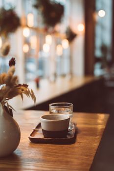 Cup of coffee espresso and glass of water on wooden table near window in cozy little cafe. Christmas winter holidays in lights of garland, cold weather, dark backdrop, defocused