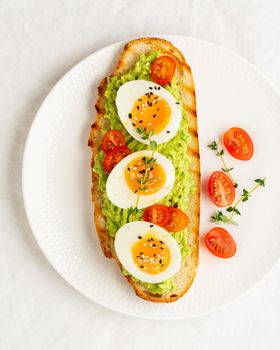 Avocado toast with toasted bread soft-boiled eggs with yellow yolk and tomatoes with herbs on white plate on light tablecloth