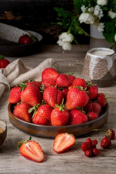 Side view of bowl with strawberry on wooden table, bottle with milk, flower. Dark still-life photo with summer berries, vertical