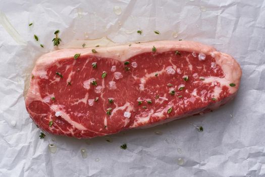 Big whole piece of raw beef meat, striploin on white parchment paper on craft background, zero waste packing. Steak with seasoning and salt. Top view, close up