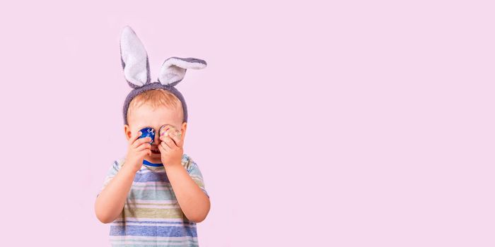 Cute baby boy in rabbit bunny ears on head closing his eyes with colored eggs on pink background. Cheerful smiling happy child. Easter holiday