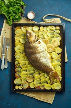 Baked carp, whole fish from the oven with sliced potatoes on a large tray on dark blue background. Traditional European Polish dish, suitable for a Christmas table