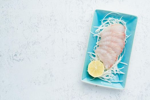 Sashimi from slices of raw white fish fillet on a blue plate on a white background. A whole Piece of fresh fish with herbs and lemon on radish. Copy space