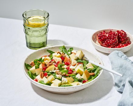 Fruits salad with nuts, balanced food, clean eating. Spinach with apples, pecans and feta, garnished with pomegranate seeds in bowl on table with white tablecloth. Hard light, shadows, side view