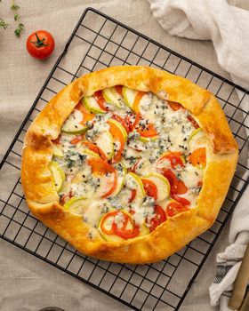 Homemade savory galette with vegetables, wheat pie with tomatoes, zucchini, blue cheese Gorgonzola. Rustic crust crostata on grid on dark linen textile tablecloth.