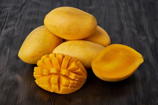 Four whole mango fruits on a wooden table and cut into slices. Large juicy bright ripe yellow fruits on a dark background, close up