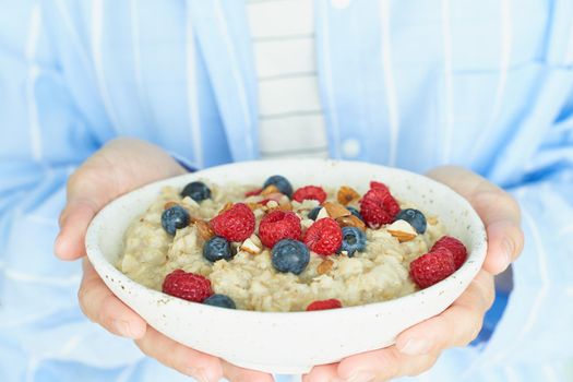 Faceless woman holds in hands breakfast, oatmeal porridge with berries and nuts, healthy food, proper nutrition. Sunshine, early morning, quiet home environment. Side view