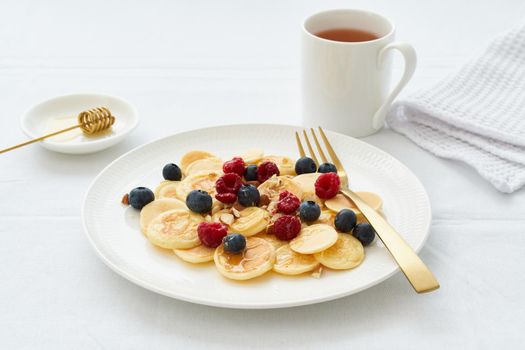 Tiny pancakes, small thin funny crumpet, children's food. Breakfast with raspberries, blueberries, honey and nuts, white tablecloth, bright morning, top view