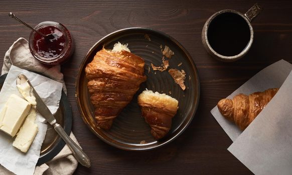 Morning breakfast with croissantand slice on plate, cup of coffee, jam and butter. Dark brown table , morning routine, slow life. Top view