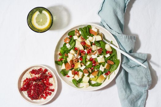 Fruits salad with nuts, balanced food, clean eating. Spinach with apples, pecans and feta, garnished with pomegranate seeds in bowl on table with white tablecloth. Hard light, shadows, top view
