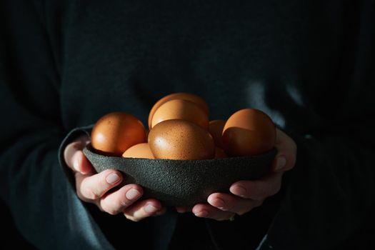 Unusual Easter on dark background. Bowl of brown eggs with hands. Darkness, rays of sunlight shine on the eggs. Concept of new life, rebirth. Rustic style. Side view, copy space