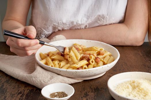 Carbonara pasta. Woman sits at table and eats pasta penne with pancetta, holding fork. Unrecognizable female in white dress. Rustic style