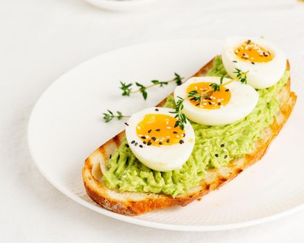 Avocado toast with toasted bread soft-boiled eggs with yellow yolk and tomatoes with herbs on white plate on light tablecloth