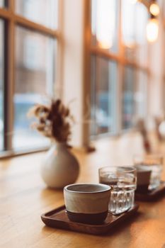 Cup of coffee espresso and glass of water on wooden table near window in cozy little cafe. Christmas winter holidays in lights of garland, cold weather, dark backdrop