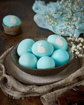 Unusual Easter on dark old background. Ceramic brown bowl with blue eggs with rabbit. Darkness, rays of sunlight shine on eggs. Concept of new life, rebirth. Rustic style. Vertical, copy space