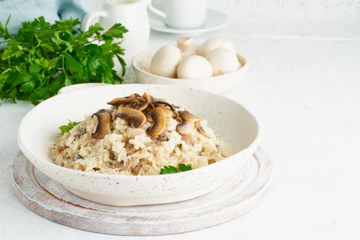 Risotto with mushrooms in plate. Rice porridge with mushrooms and parsley. White table, spoons, mushrooms. Hot dish, italian cuisine, side view, copy space