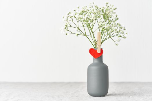 Valentine's Day. Delicate white flowers in a vase. Red felt heart - symbol of lovers. Light white gentle pastel background. Scandinavian minimalism. Side view, copy space