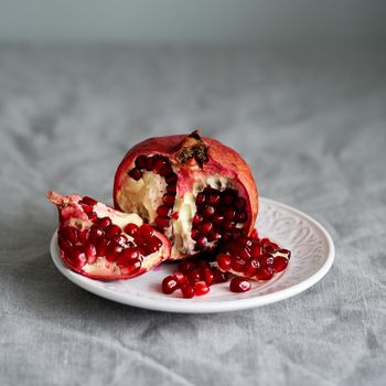 Still life with broken open pomegranate and seeds on plate on table covered with crumpled gray tablecloth in natural light from window. Lifestyle photo with bright red center on neutral backdrop