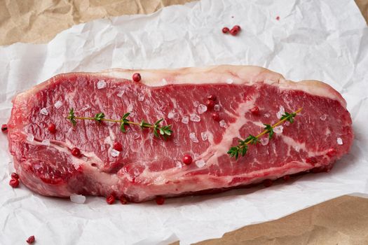 Big whole piece of raw beef meat, striploin on white parchment paper on craft background, zero waste packing. Seasoning steak with salt. Side view