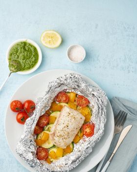 Foil pack dinner with white fish. Oven baked fillet of cod, pike perch with vegetables and pesto sauce. Diet food, keto diet, Mediterranean cuisine. Blue background. Top view, vertical, copy space