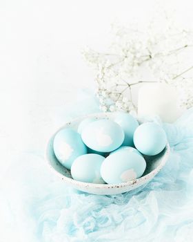 Easter. Holiday. Light white background, gentle pastel colors. Blue eggs with image of rabbit in basket. Flowers in background, side view, vertical