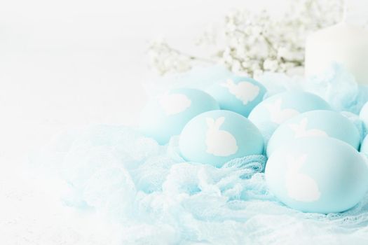 Easter. Holiday. Light white background, gentle pastel colors. Blue eggs with image of rabbit in basket. Flowers in background, side view, copy space