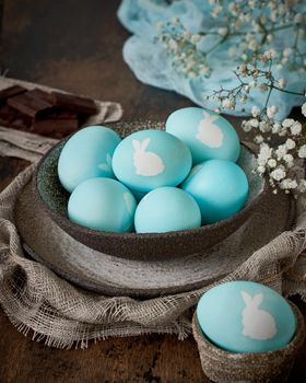 Unusual Easter on dark old background. Ceramic brown bowl with blue eggs with rabbit. Darkness, rays of sunlight shine on eggs. Concept of new life, rebirth. Rustic style. Vertical, copy space