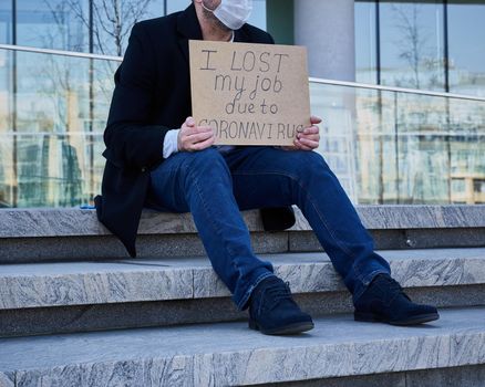 Job loss due to COVID-19 virus pandemic concept. Unrecognizable man holds sign I lost my job because of coronavirus. Male sitting on stairs of building, former place of work in business center