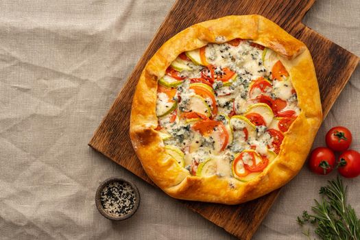 Homemade savory galette with vegetables, wheat pie with tomatoes, zucchini, blue cheese Gorgonzola. Rustic crust crostata on dark linen textile tablecloth.