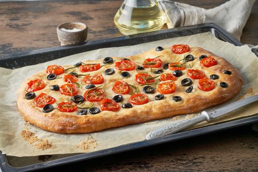Focaccia, pizza, italian flat bread with tomatoes, olives and rosemary on tray on dark wooden rustic table, side view, close up