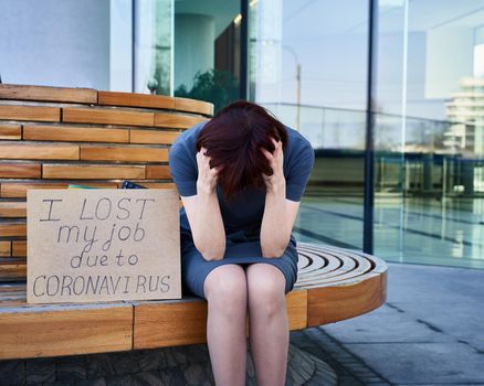 Woman holds sign saying I lost my job because of coronavirus. Concept of job loss due to the COVID-19 virus pandemic. Female with closed eyes stands against background of business cente