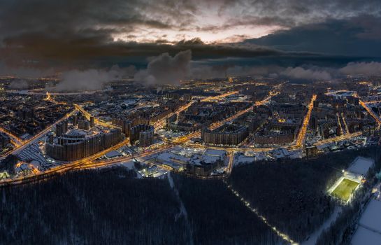 Aerial winter cityscape of St. Petersburg - Russia at dusk, drone flies over huge housing estates and public park, construction cranes, night illumination, clouds float below drone. High quality photo