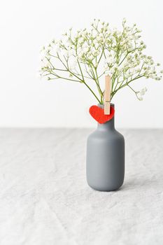 Valentine's Day. Delicate white flowers in a vase. Red felt heart - symbol of lovers. Light white gentle pastel background. Scandinavian minimalism. Side view, copy space, vertical