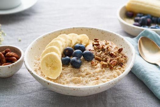 Whole oatmeal, large bowl of porridge with banana, blueberries, nuts for breakfast, morning meal. Side view, close up, gray table. Vegan tasty healthy diet.