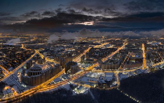 Aerial winter cityscape of St. Petersburg - Russia at dusk, drone flies over huge housing estates and public park, construction cranes, night illumination, clouds float below drone. High quality photo