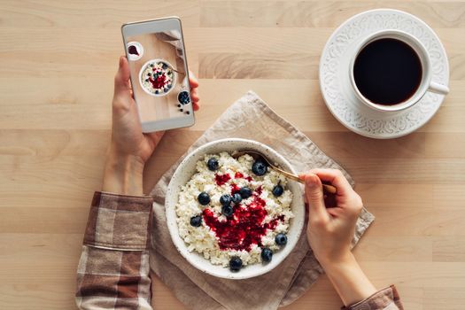 Blogger taking photos of food, shooting Breakfast on mobile phone, bowl of homemade curd with jam, raspberries, blueberries and cup of coffee